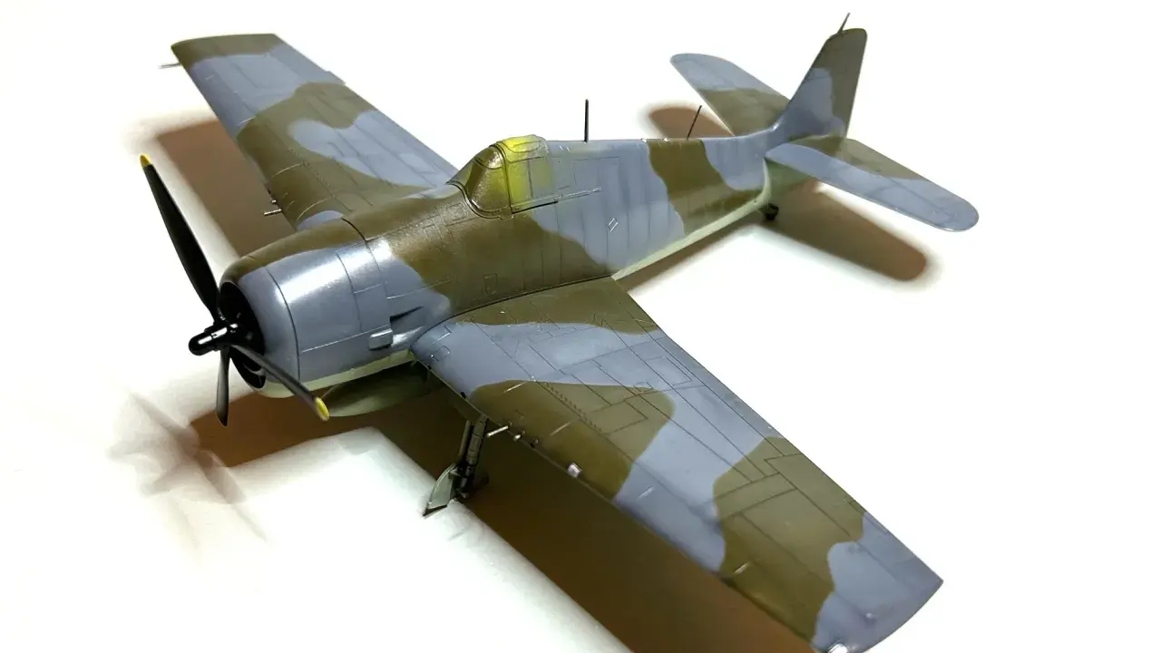 Ein Modellflugzeug, ein Hellcat-Flugzeug, mit Tarnmuster in Grau und Olivgrün, steht auf weißem Hintergrund.