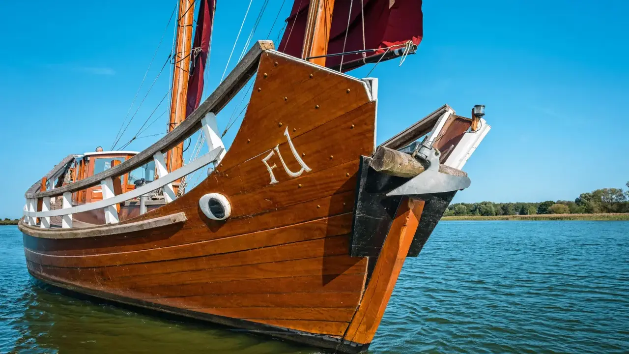 Ein hölzernes Dschunke Schiff gleitet auf ruhigem Wasser unter blauem Himmel. Die Segel sind teilweise gesetzt.
