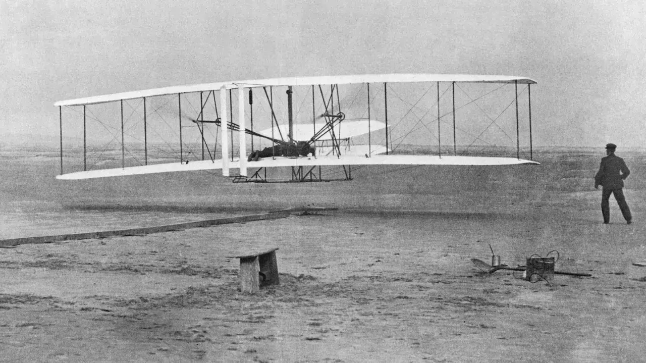 Wright Flyer mit Doppelruder hebt ab, Pilot liegt auf dem Flügel. Ein Mann beobachtet den historischen Flug.