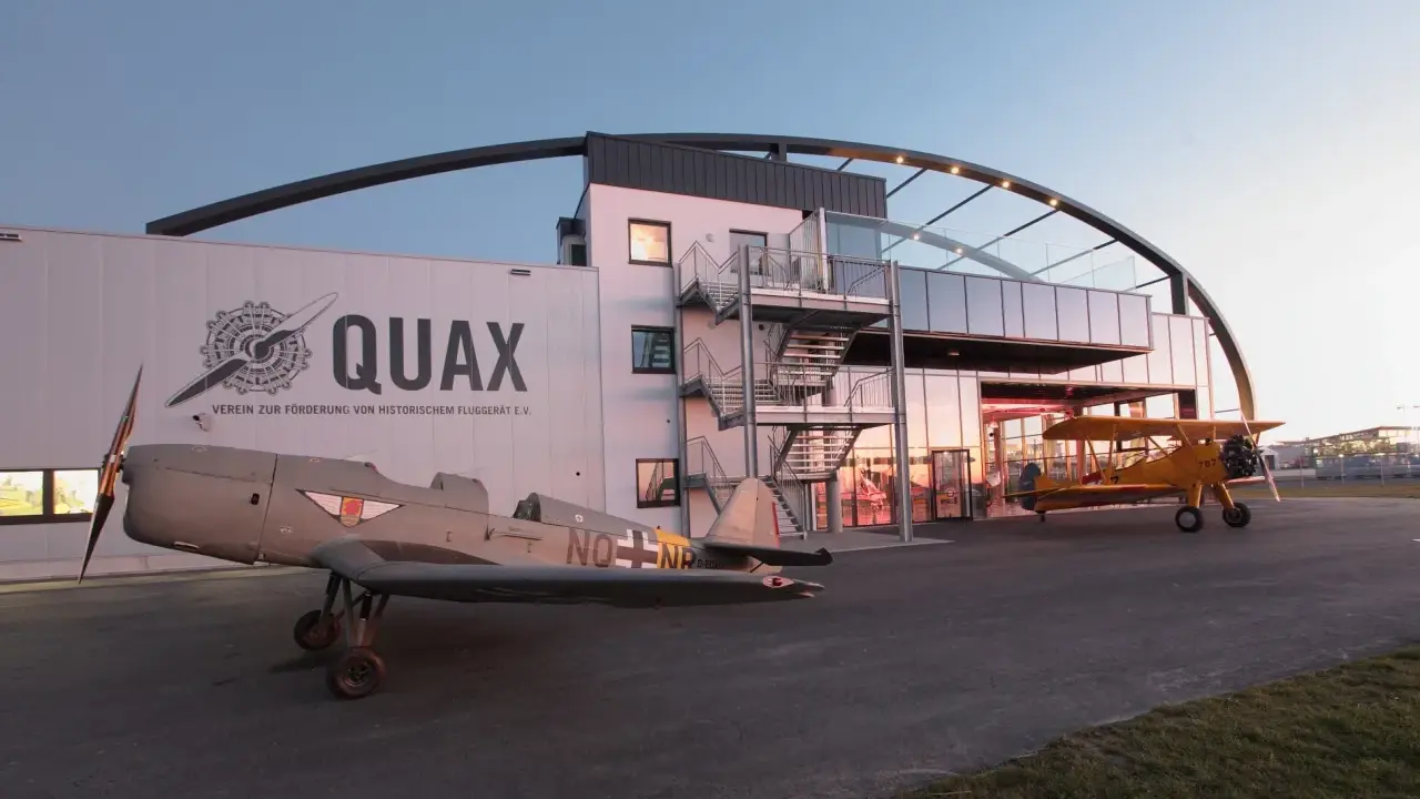 Zwei historische Flugzeuge stehen vor dem Quax Hangar. Ein graues Propellerflugzeug im Vordergrund, ein gelbes Doppeldeckerflugzeug daneben.