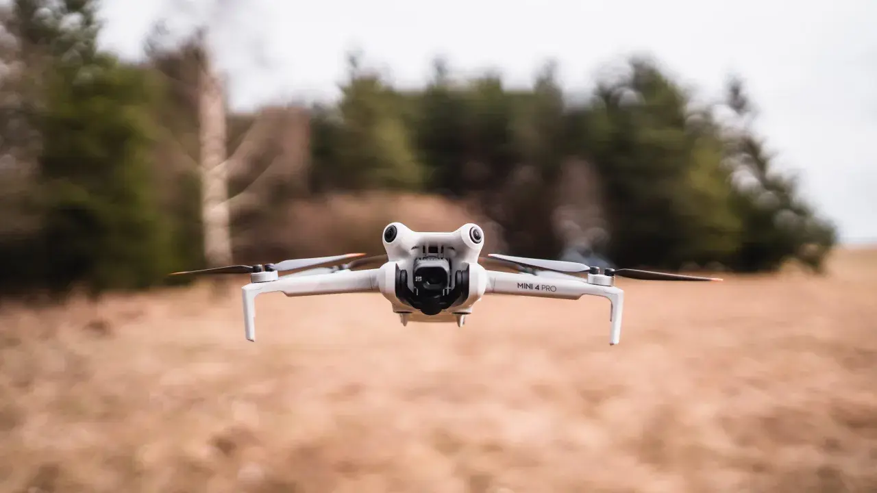 Eine weiße Drohne A3, Modell Mini 4 Pro, schwebt über trockenem Gras. Im Hintergrund verschwimmen Bäume und eine bewölkte Himmel.