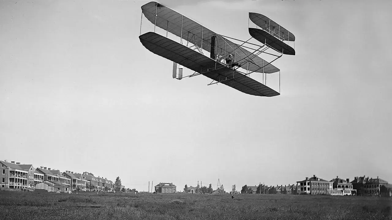 Das Kitty Hawk Flugzeug fliegt über ein Feld mit Gebäuden.