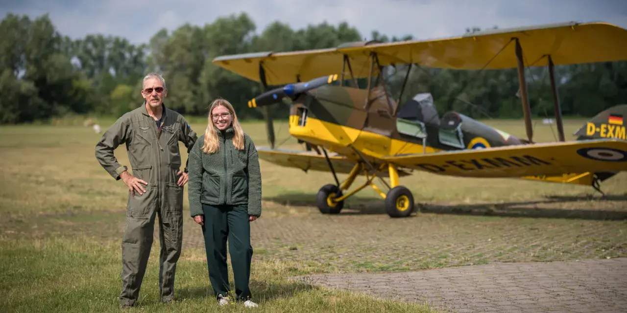 Mann in Fliegerkluft und junge Frau stehen neben gelbem Doppeldeckerflugzeug. Das Flugzeug erinnert an einen Tiger Moth.