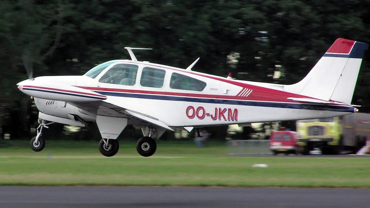 Eine weiße Beechcraft Bonanza mit roten und blauen Streifen hebt vom Gras ab.