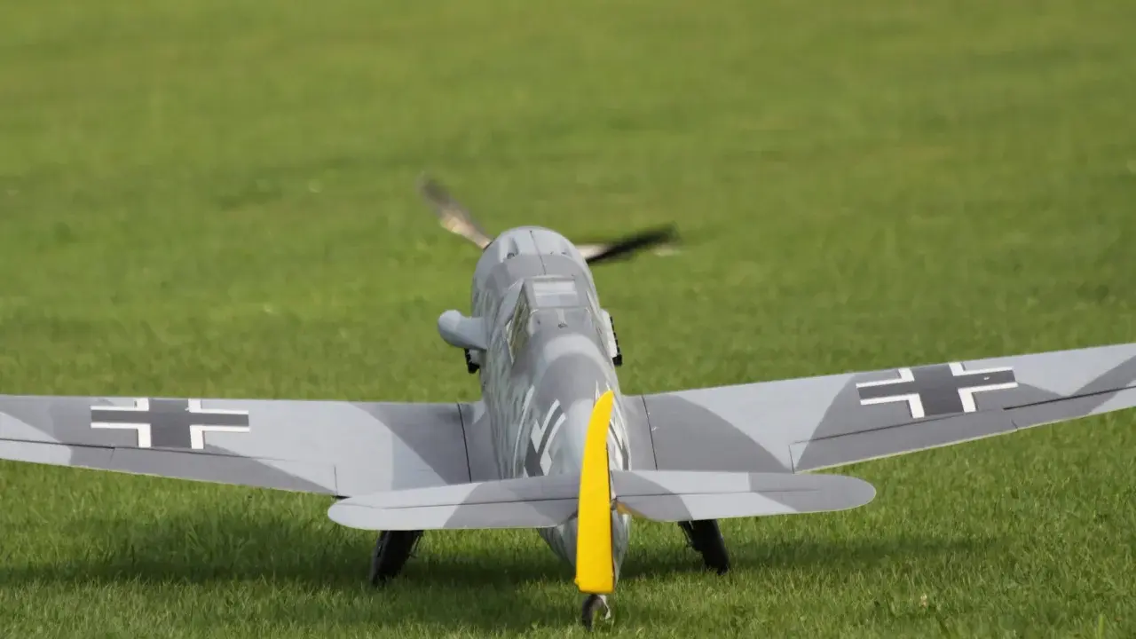 Modellflugzeug einer Me109 mit Tarnmuster und gelbem Seitenleitwerk auf einer grünen Wiese.