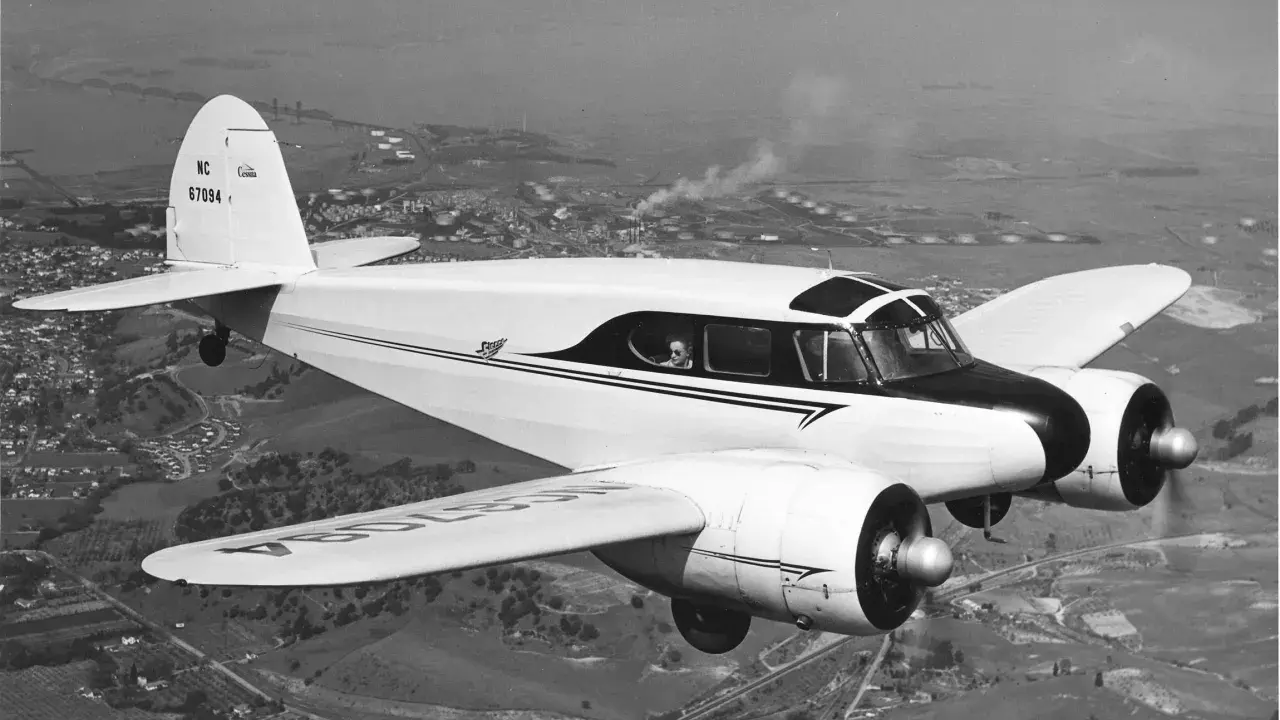 Ein zweimotoriges Flugzeug, ein klassisches Cessna-Modell, fliegt über eine ländliche Landschaft mit einer Stadt und Industrieanlagen im Hintergrund.