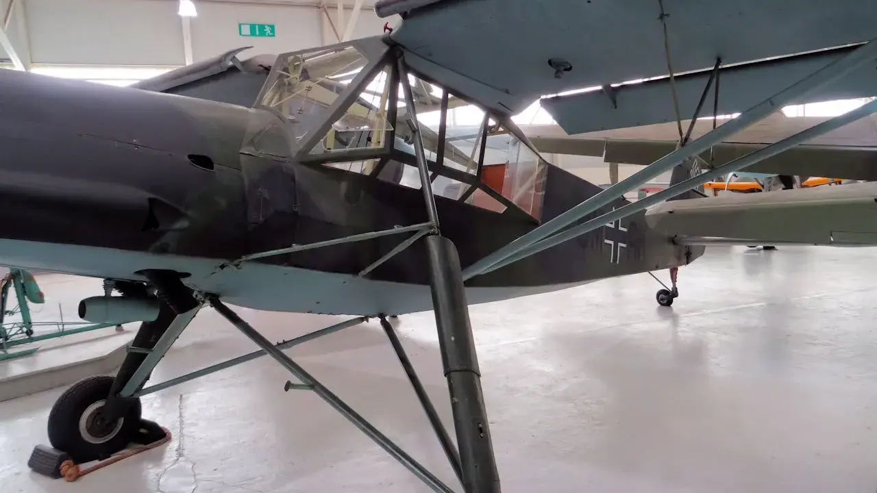Ein altes Flugzeug, ein Storch, steht in einer Halle. Das Flugzeug hat eine Tarnlackierung und ein Kreuz auf dem Heck.
