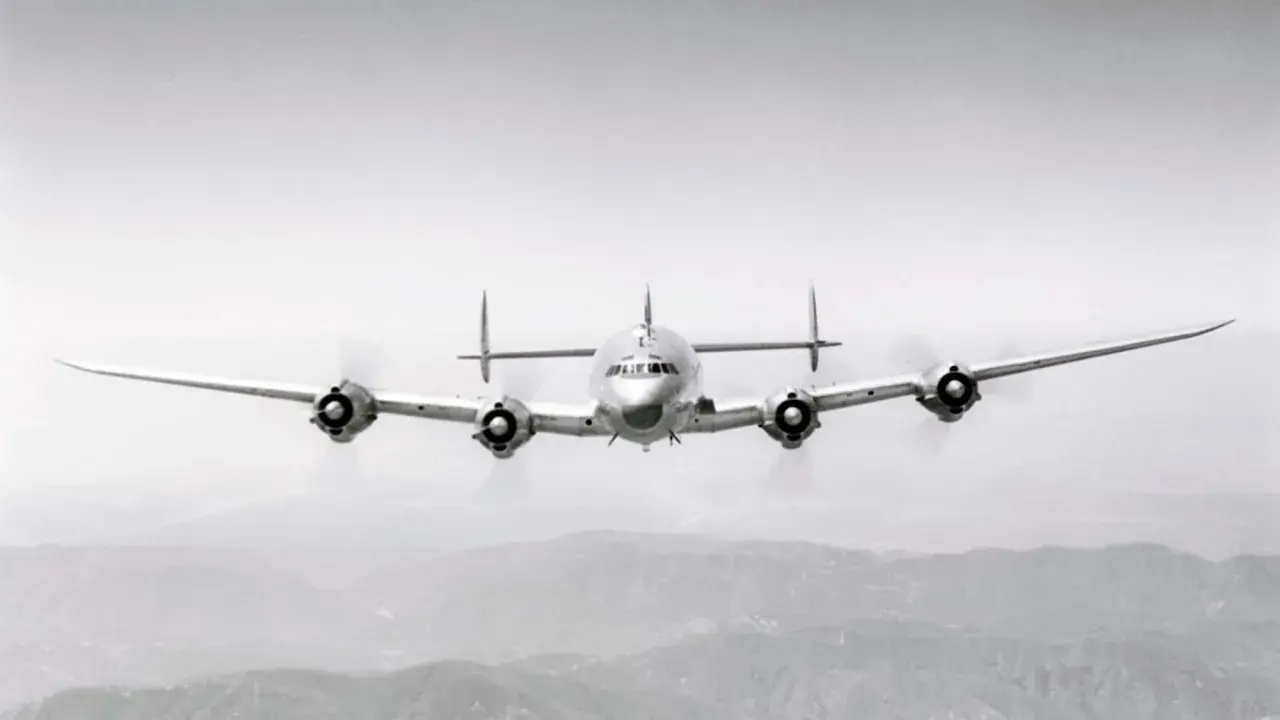 Ein vier-motoriger Propellerflieger mit markantem Leitwerk fliegt über bergiger Landschaft.