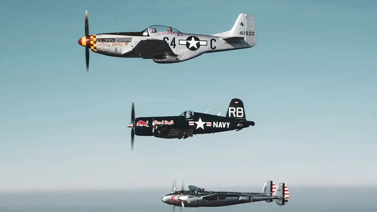 Drei historische Flugzeuge im Flug: eine silberne P-51 Mustang, ein schwarzer Red Bull Flieger (Corsair) und eine P-38 Lightning.