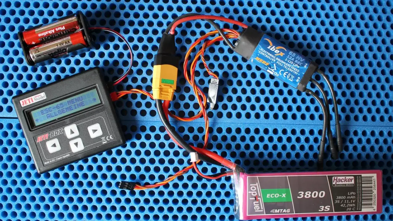 Jeti Box mit ESC-65-Menü, ibex 65 HV ESC, Hacker LiPo-Akku und Batterien auf blauem Untergrund.