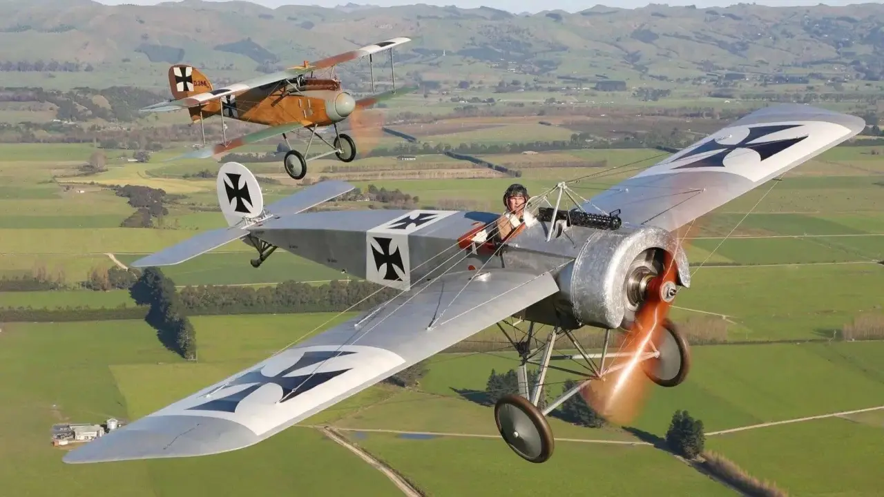 Zwei historische Fokker Eindecker fliegen über grüne Felder. Ein Pilot im Vordergrund, der andere im Hintergrund.