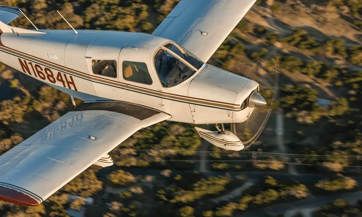 Eine Piper Cherokee 140 fliegt über eine bewaldete Landschaft.