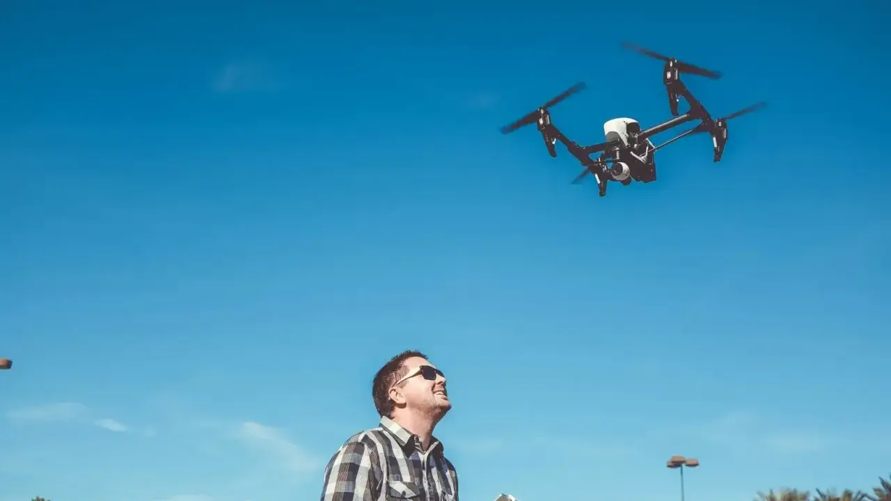 Ein **Drohnenpilot** steuert mit einer Fernbedienung einen Quadrocopter am blauen Himmel.