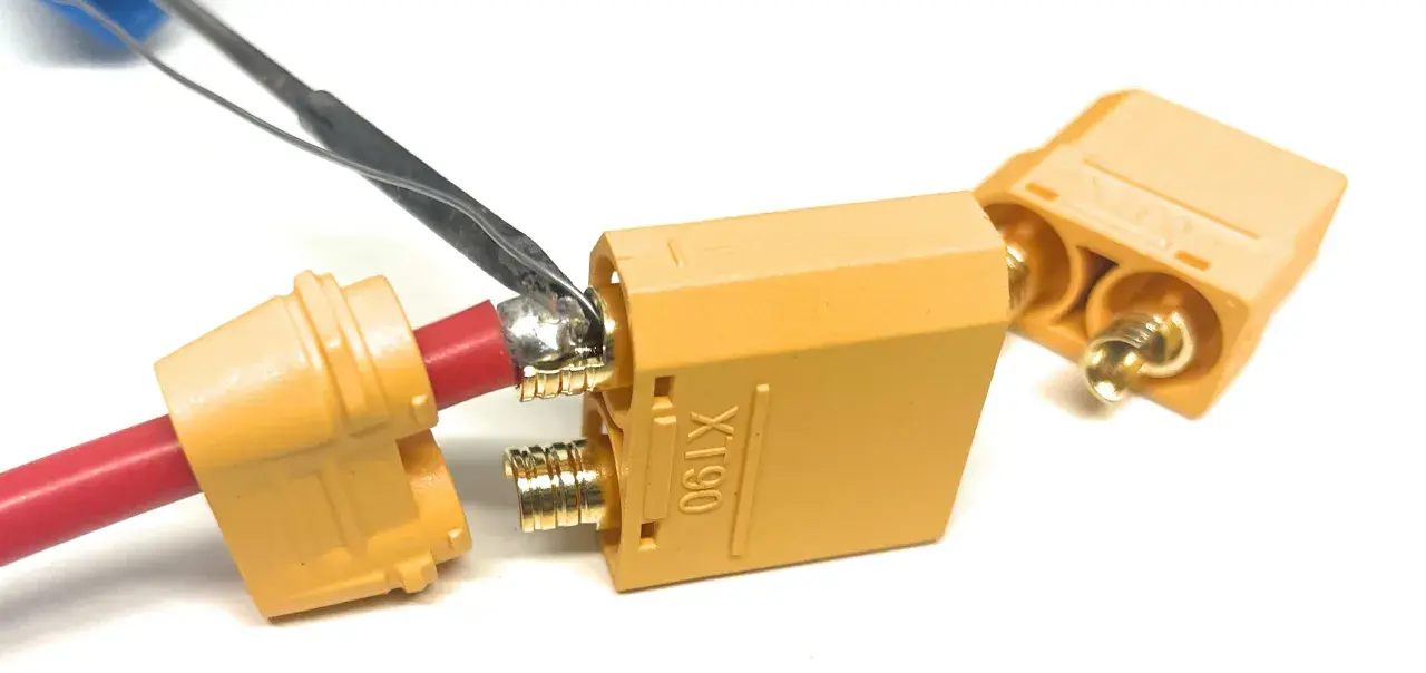 Löten eines roten Kabels an einen goldenen XT90-Stecker. Ein weiterer Stecker liegt daneben.
