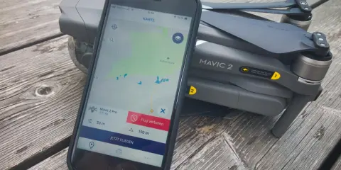 Eine Drohne liegt neben einem Smartphone, auf dem eine Drohnen-App mit Karte und Flugverboten angezeigt wird.
