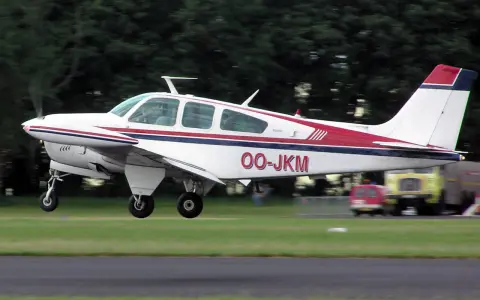 Eine weiße Beechcraft Bonanza mit roten und blauen Streifen hebt vom Gras ab.