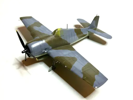 Ein Modellflugzeug, ein Hellcat-Flugzeug, mit Tarnmuster in Grau und Olivgrün, steht auf weißem Hintergrund.