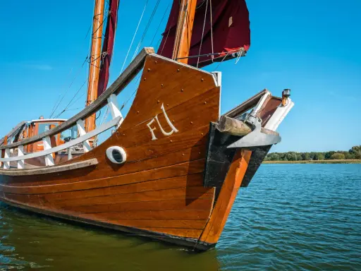 Ein hölzernes Dschunke Schiff gleitet auf ruhigem Wasser unter blauem Himmel. Die Segel sind teilweise gesetzt.