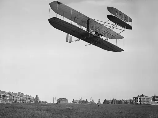 Das Kitty Hawk Flugzeug fliegt über ein Feld mit Gebäuden.