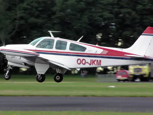 Eine weiße Beechcraft Bonanza mit roten und blauen Streifen hebt vom Gras ab.