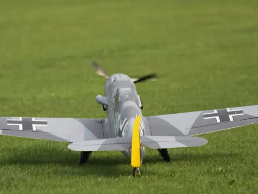 Modellflugzeug einer Me109 mit Tarnmuster und gelbem Seitenleitwerk auf einer grünen Wiese.