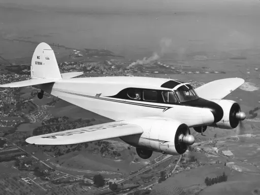 Ein zweimotoriges Flugzeug, ein klassisches Cessna-Modell, fliegt über eine ländliche Landschaft mit einer Stadt und Industrieanlagen im Hintergrund.