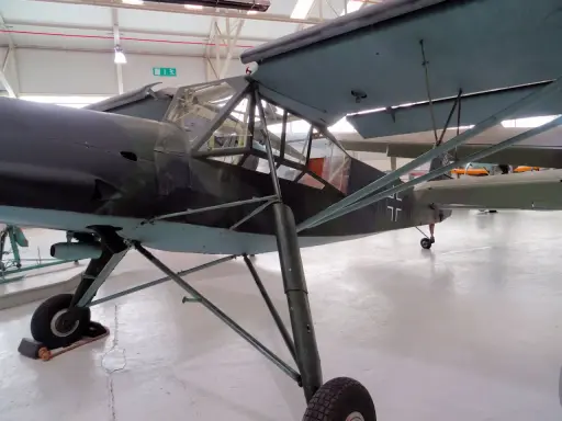 Ein altes Flugzeug, ein Storch, steht in einer Halle. Das Flugzeug hat eine Tarnlackierung und ein Kreuz auf dem Heck.