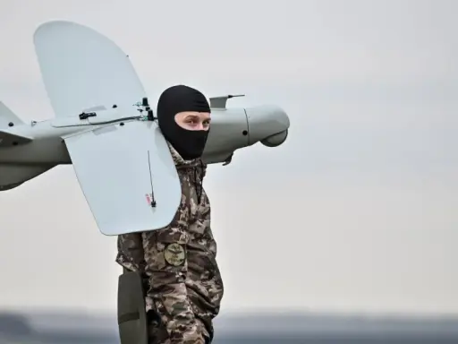 Mann in Tarnkleidung und schwarzer Sturmhaube trägt eine Drohne. Seit wann gibt es Drohnen? Die Geschichte reicht weit zurück.