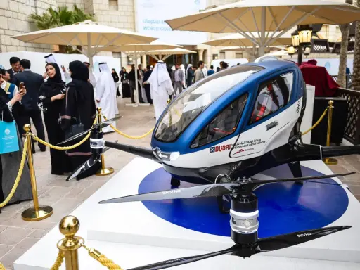 Ein futuristisches Flugtaxi, ein Dubai Drohne, steht auf einer Ausstellungsplattform. Menschen in traditioneller Kleidung betrachten das Gefährt.