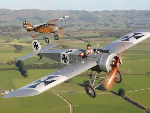 Zwei historische Fokker Eindecker fliegen über grüne Felder. Ein Pilot im Vordergrund, der andere im Hintergrund.