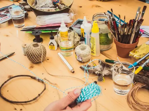 Bastelmaterialien auf Holztisch, darunter Schnur, Federn, Pinsel und eine kleine Flasche uhu super glue.