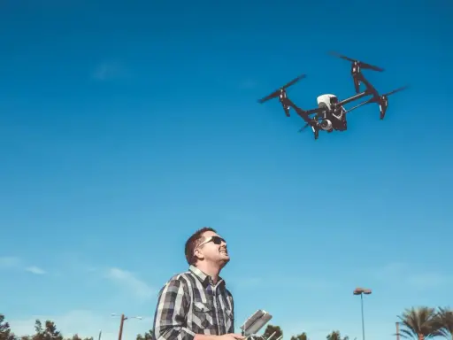 Ein **Drohnenpilot** steuert mit einer Fernbedienung einen Quadrocopter am blauen Himmel.