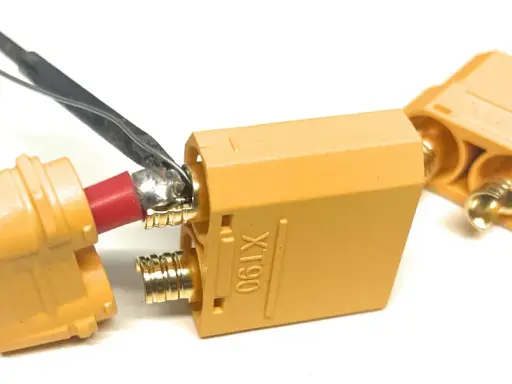Löten eines roten Kabels an einen goldenen XT90-Stecker. Ein weiterer Stecker liegt daneben.