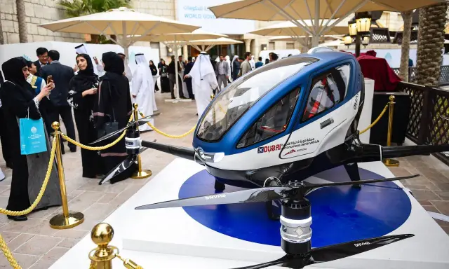 Ein futuristisches Flugtaxi, ein Dubai Drohne, steht auf einer Ausstellungsplattform. Menschen in traditioneller Kleidung betrachten das Gefährt.