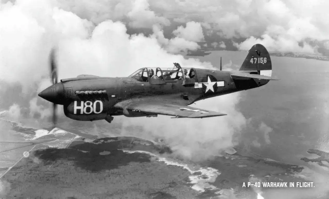 Eine P-40 Warhawk mit der Kennung H80 fliegt über eine Küstenlandschaft.