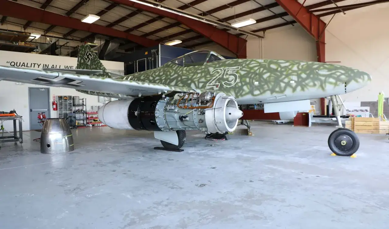 Messerschmitt Jet mit Tarnmuster und freiliegendem Triebwerk in einer Werkstatthalle.