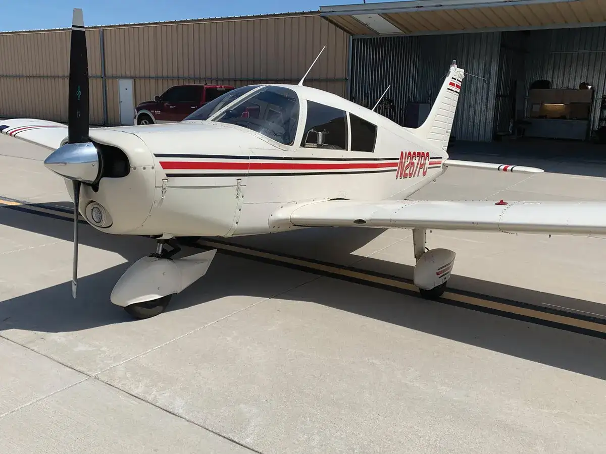 Weiße Piper Cherokee 140 mit roten und schwarzen Streifen steht auf dem Rollfeld vor einer Flugzeughalle.