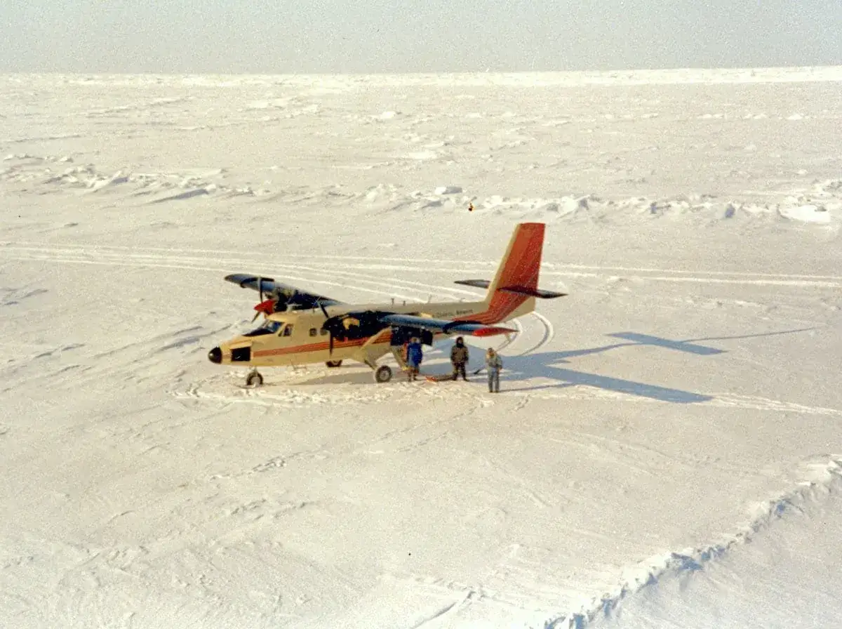 Ein DHC-6 Twin Otter steht auf einer eisigen, schneebedeckten Fläche. Drei Personen stehen neben dem Flugzeug.