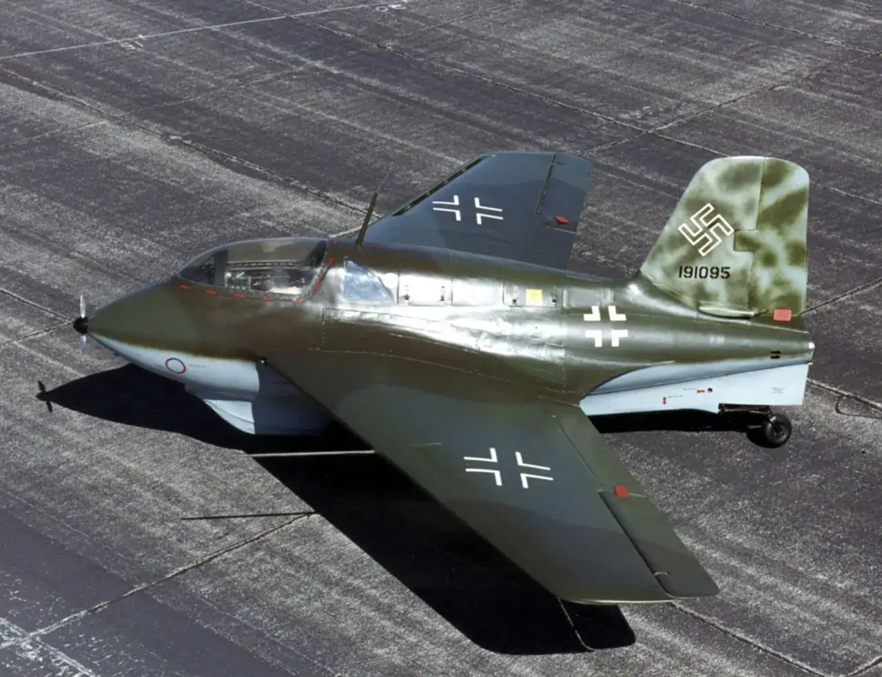 Ein Me 163 Komet, ein Raketenjäger, steht auf einem Flugplatz. Seine Flügel und das Leitwerk tragen weiße Balkenkreuze.