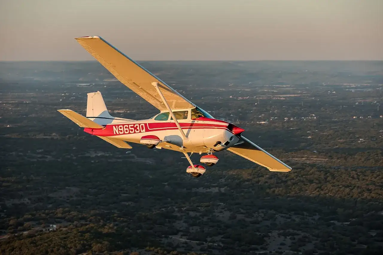 Eine wei&szlig;e und rote Cessna Skyhawk fliegt &uuml;ber eine bewaldete Landschaft im goldenen Licht des Sonnenuntergangs.