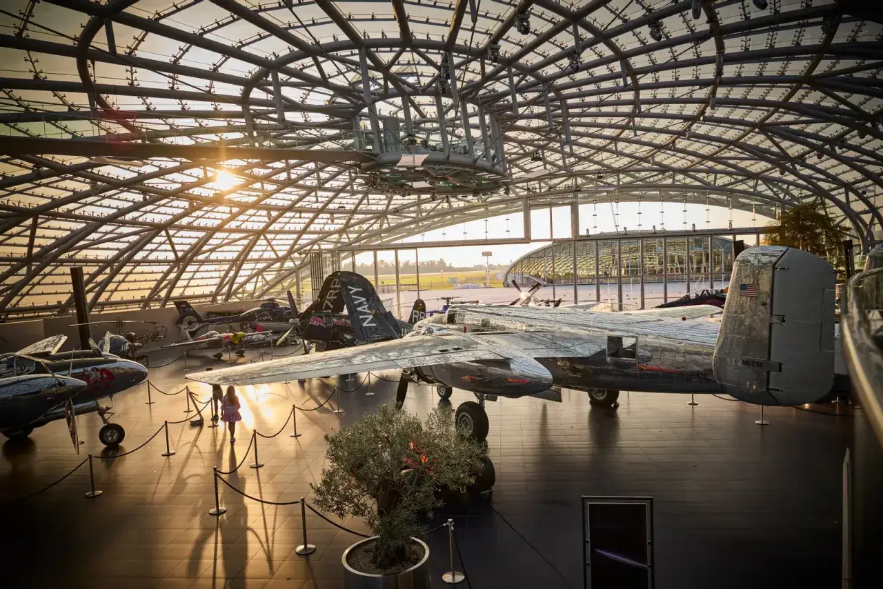Sonnenlicht fällt durch das Glasdach auf historische Flugzeuge, darunter ein glänzender Red Bull Flieger.