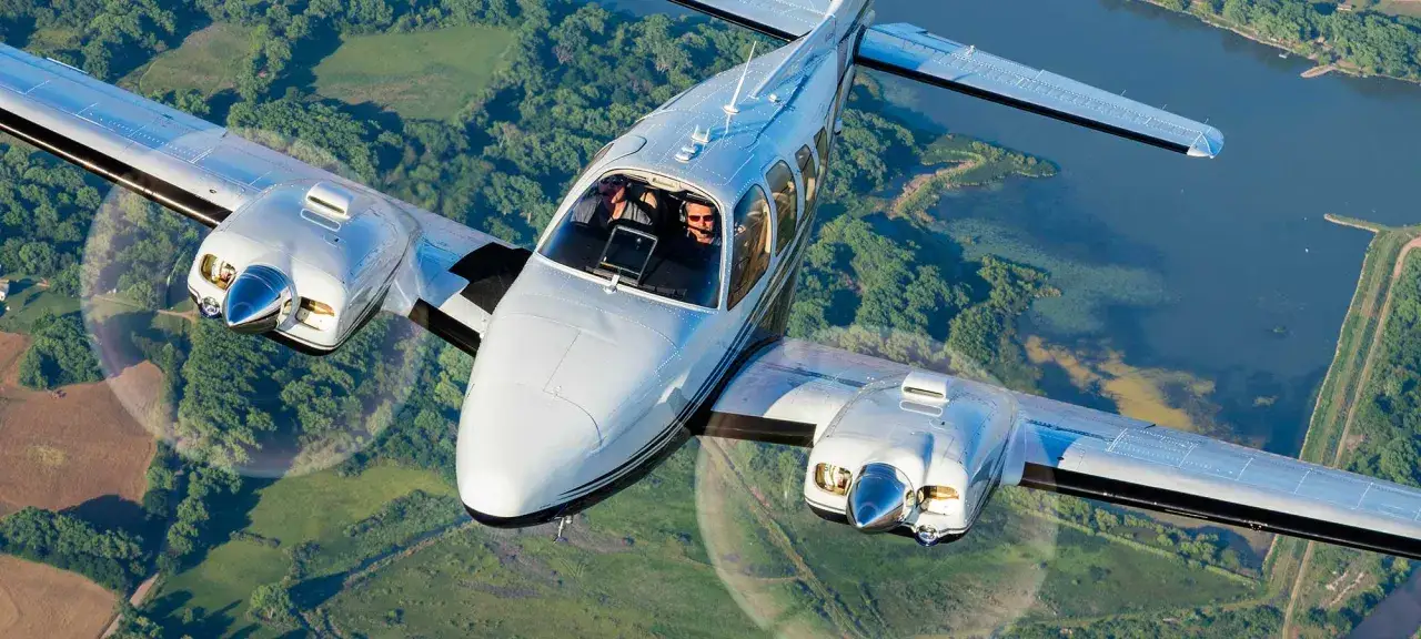 Zwei Piloten steuern eine glänzende Beechcraft Baron über grüne Wälder und einen blauen See.