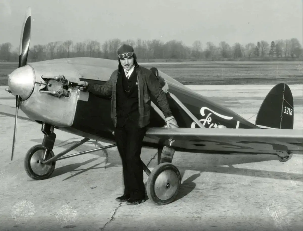Ein Pilot in Fliegerkleidung steht neben einem Ford Flugzeug. Das Flugzeug hat einen Propeller und R&auml;der.