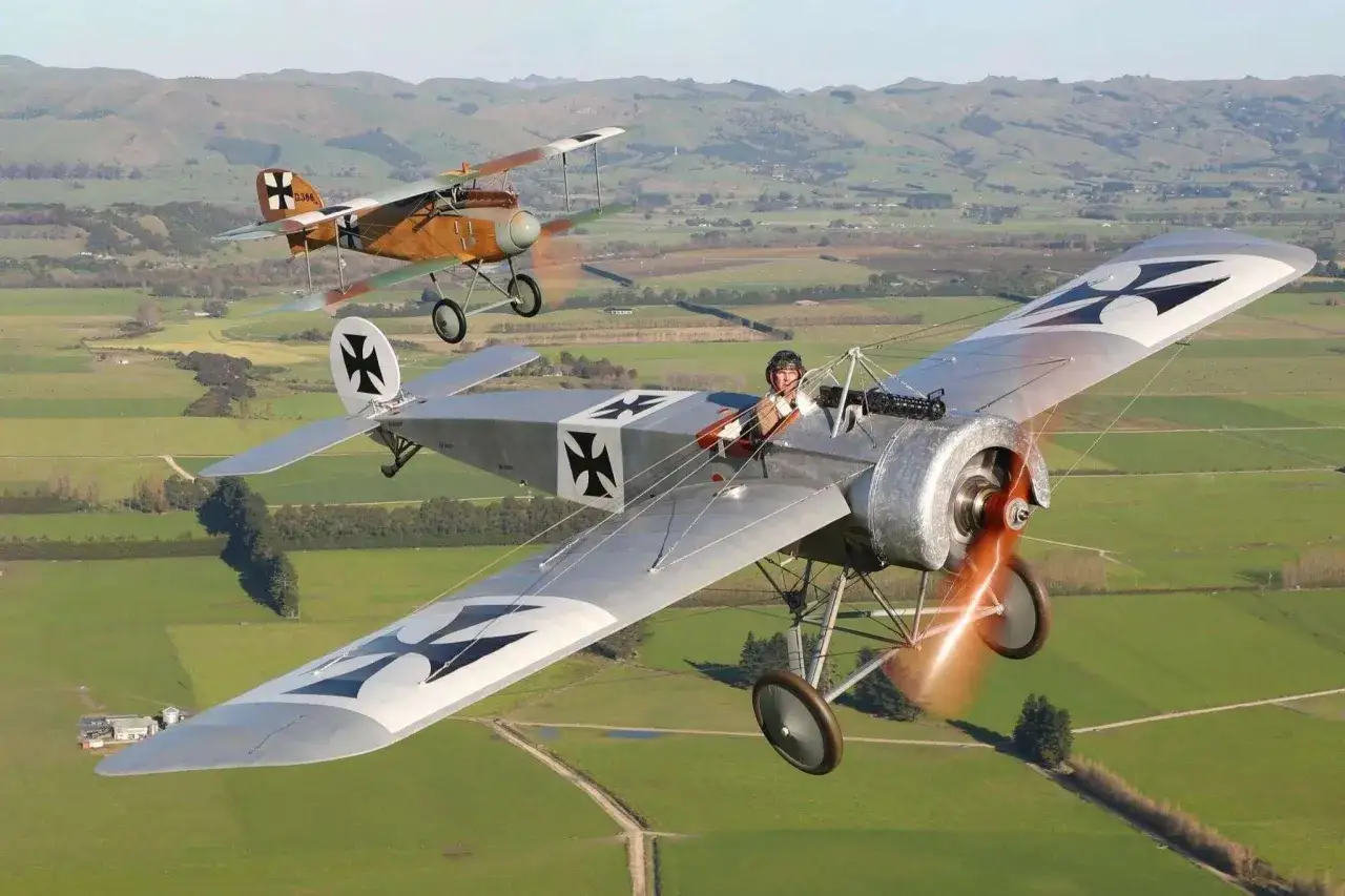 Zwei historische Fokker Eindecker fliegen über grüne Felder. Ein Pilot im Vordergrund winkt.