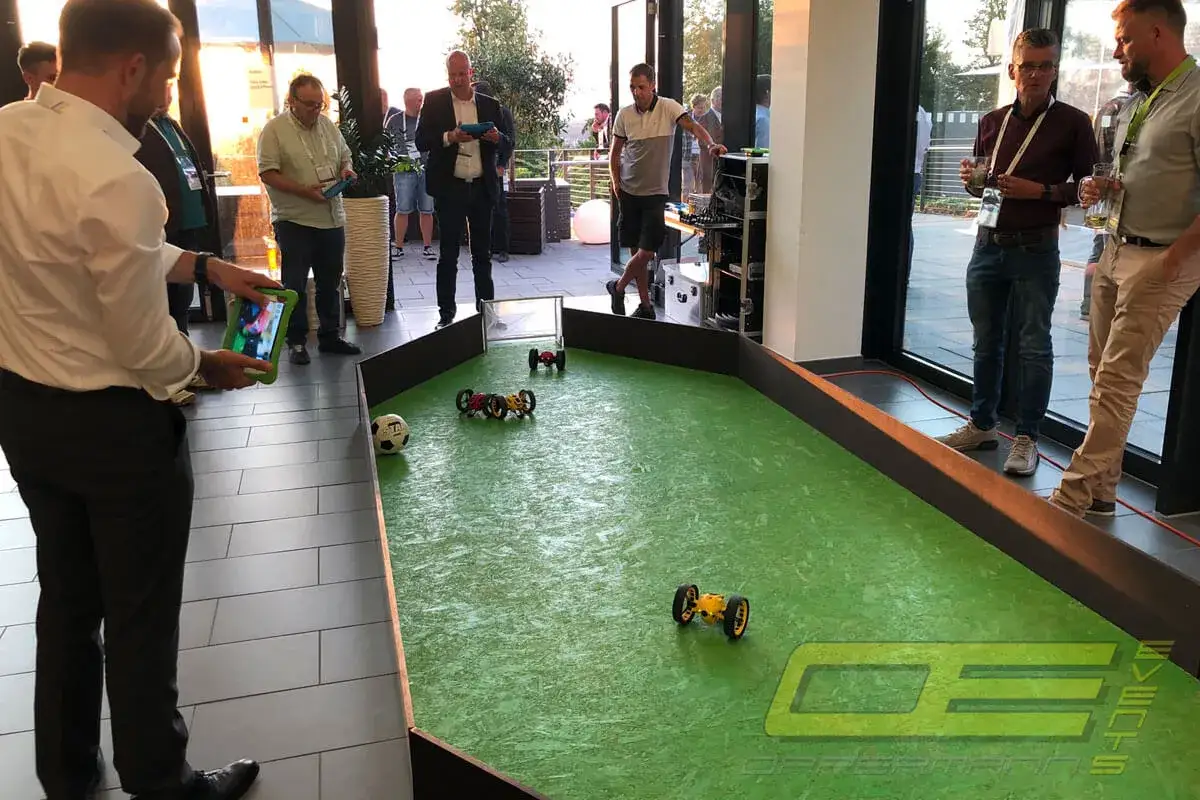 Männer steuern ferngesteuerte Autos auf einem grünen Spielfeld, ein Fußball liegt daneben. Ein spannender Drohnen Sport-Moment.