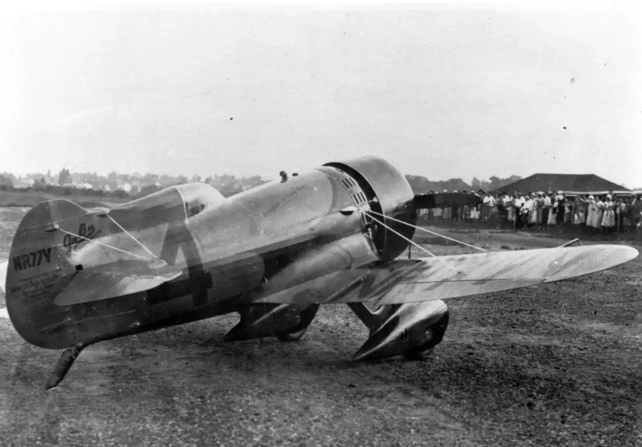Ein altes Flugzeug, eine Gee Bee Rennmaschine, steht auf einem Feld. Eine Menschenmenge beobachtet das historische Fluggerät.