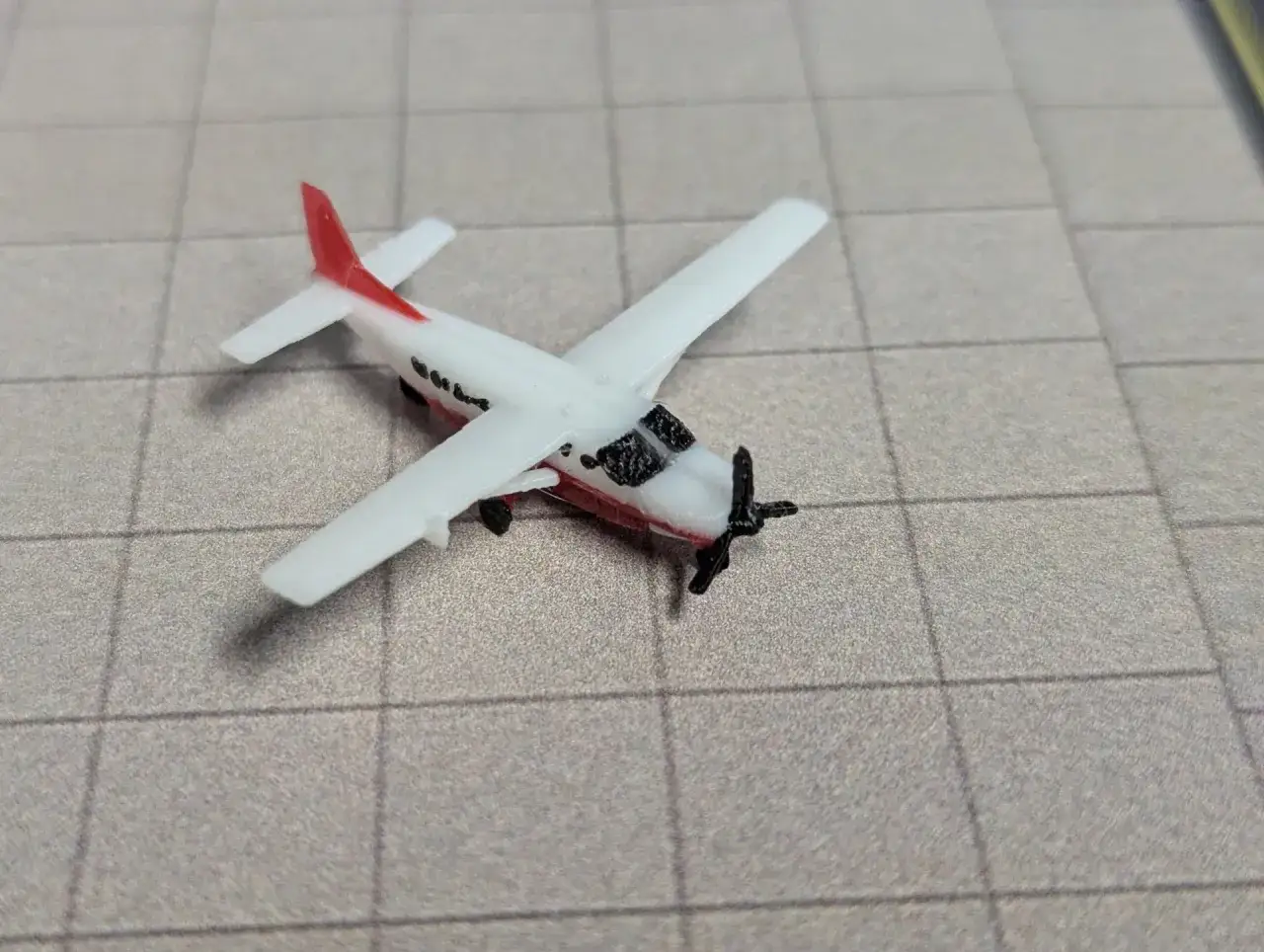 Kleines Modellflugzeug, eine weiße Cessna 208 Caravan mit rotem Leitwerk, auf kariertem Untergrund.