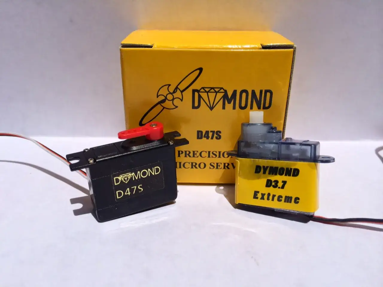 Zwei Servos, ein schwarzer DYMOND D47S und ein gelber D3.7 Extreme, stehen vor einer gelben DYMOND-Box.
